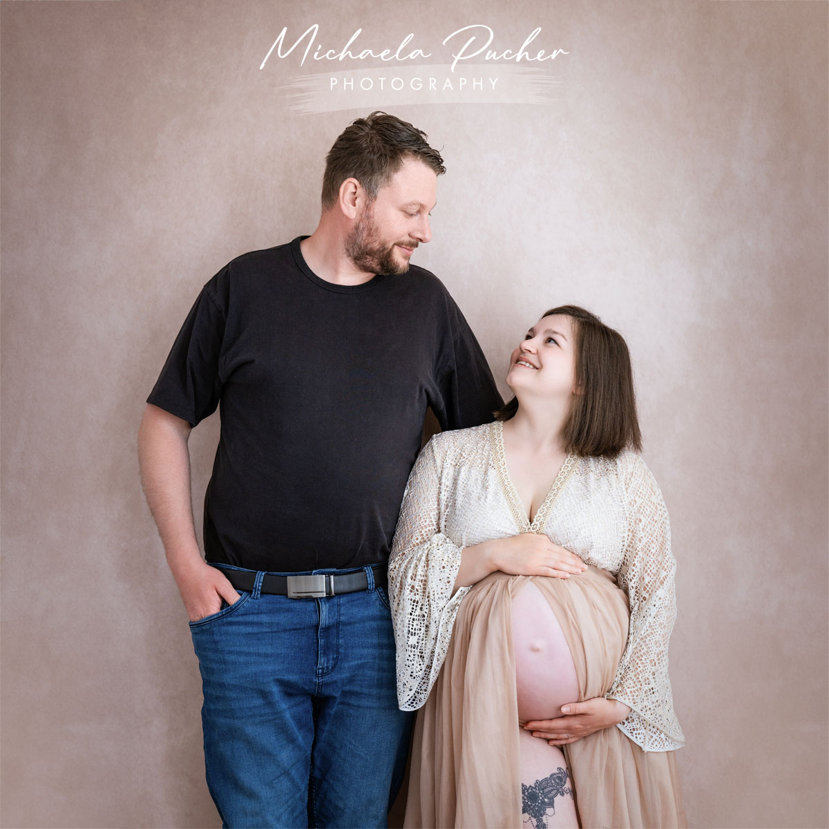 Schwangere Frau steht lächelnd neben ihrem Partner, beide blicken sich liebevoll an; die Frau hält ihren Babybauch, studiofotografiert vor einem hellen, neutralen Hintergrund.